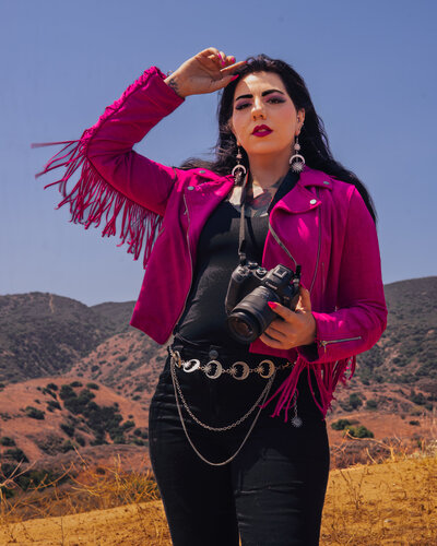 Close-up branding portrait of a Utah photographer in a bold pink fringe jacket holding a Canon camera, photographed in the desert hills near Salt Lake City. Editorial, modern branding imagery by Moonlit Photo.