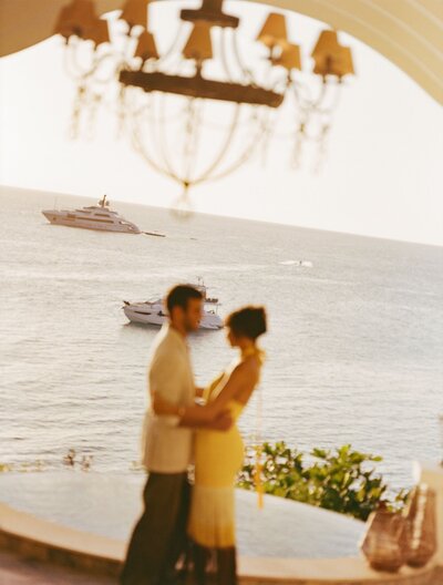 A couple stands closely under a large chandelier, gazing at each other, with the ocean and yachts in the background. The scene is softly blurred, creating a dreamy atmosphere.