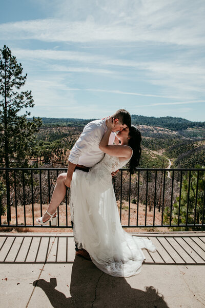 A black-and-white photo of a couple during their Lake Tahoe elopement. The man lifts the woman in his arms as they laugh, her hair blowing in the wind. They stand at the water’s edge, with large boulders and distant mountains framing the scenic lake setting.