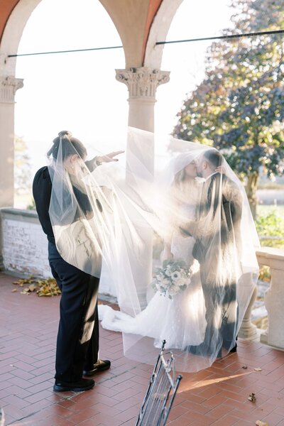 Behind-the-scenes moment of a photographer adjusting a long veil as a bride and groom share a kiss under it, framed by stone architecture and glowing sunlight.