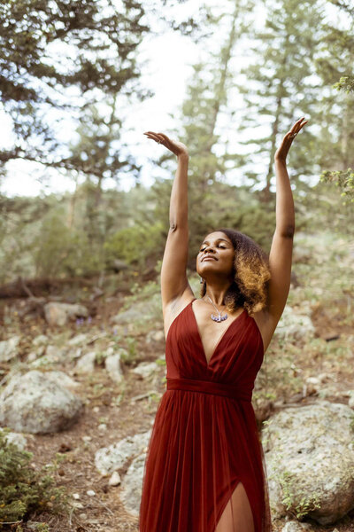 woman with a flower crown and a red dress standing in the forest with both hands and arms toward the sky