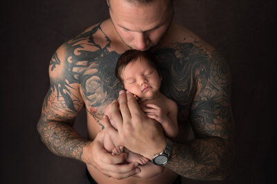 Tattooed father tenderly cradling his newborn baby against his chest during a luxury Dallas newborn photography session. The baby sleeps peacefully, highlighting the strength and love of fatherhood.