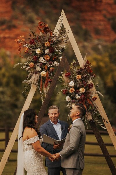 A wedding ceremony in Sedona, Arizona, where the couple exchanges vows under a beautifully decorated triangular wooden arch. The officiant stands between the marriers, who hold hands and smile at each other, with red rock formations in the background.