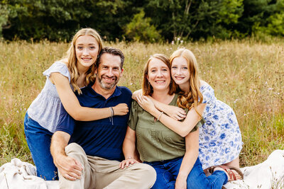 mother and father sitting together as their two daughters hug them from behind in a field near Milwaukee