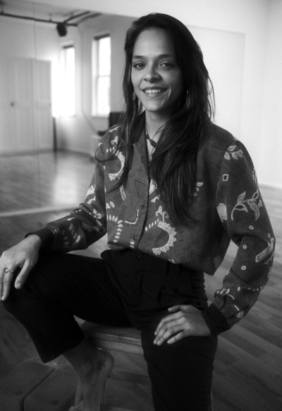 Black and white photo of woman with short hair smiling vibrantly. She is sitting in a chair in a button down shirt, holding one leg up.