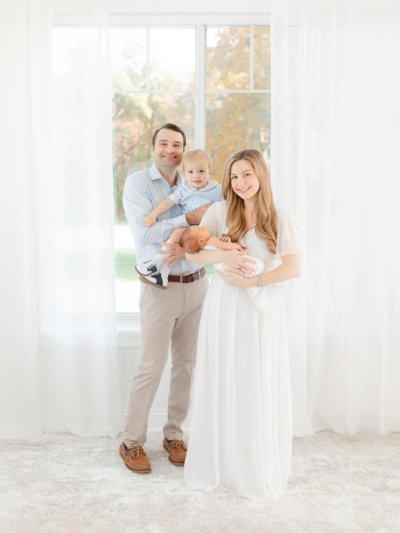 family with toddler and newborn smiles at the camera during boston newborn photoshoot