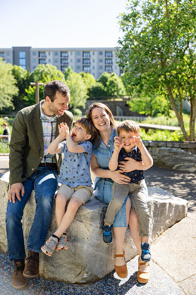 A man and woman sit on a rock with two children, enjoying a sunny day outdoors.