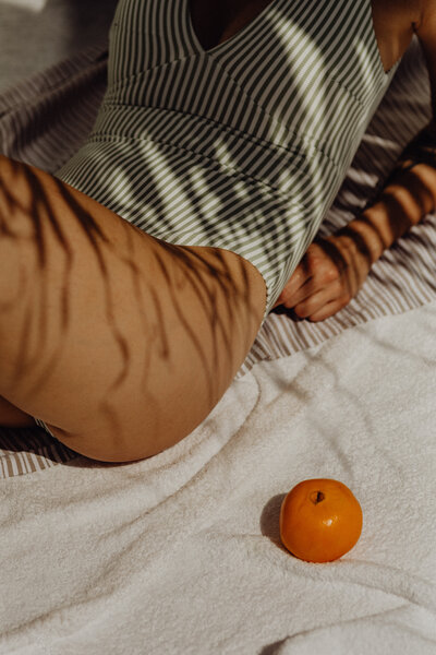 A golden-tanned woman lounges in a navy and white vertically striped one-piece swimsuit, resting on her elbows with her legs bent as if sunbathing. Leaf shadows dance across her skin, creating a dreamy summer aesthetic. Captured for Sunny Days Swimwear’s branding, this image embodies effortless beachside luxury.