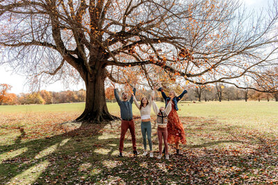 Connecticut family photographer documents joyful family of four throwing autumn leaves under large tree in fall park setting
