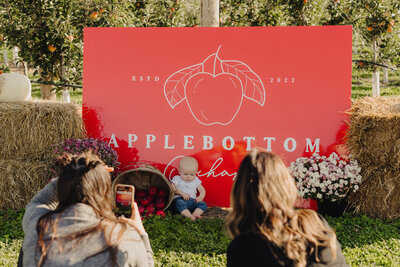 Baby sitting in front of Applebottom Orchards red sign with fall mums and haybales.