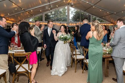 The newlywed couple enters their Philadelphia wedding reception hand in hand, beaming as guests rise to cheer them on. The bride’s lace wedding gown catches the glow of string lights draped across the venue’s tented ceiling, while the groom’s classic black suit perfectly complements the elegant rustic decor