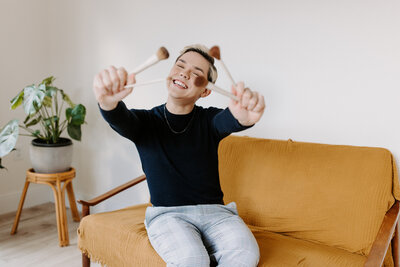 person sitting on a couch smiling while holding out paint brushes in both hands