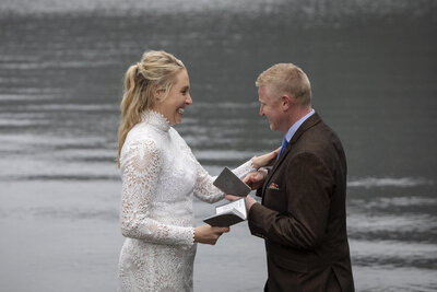 Bride and groom walk barefoot through the shallow waters of a rocky Alaskan shoreline at sunset, with rugged snow-capped mountains in the background. The groom wears rolled-up pants and suspenders, while the bride lifts her lace wedding dress, embracing the adventurous spirit of an Alaska elopement.