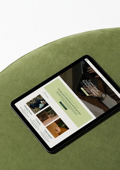 Tablet mockup featuring the Big Bear Showit website template open on screen. The homepage includes soft sage-green sections with elegant serif text, showcasing photos of cozy interiors and a minimalist layout. The tablet sits on a round green velvet surface photographed from above.
