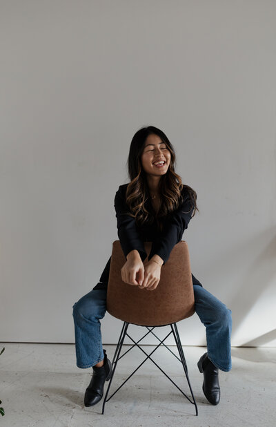 A woman wearing a black sweater and blue jeans sits backwards on a brown chair, smiling with eyes closed against a plain white wall. With long hair catching the indoor sunlight, she radiates a joy captured perfectly by a documentary photographer in New England.