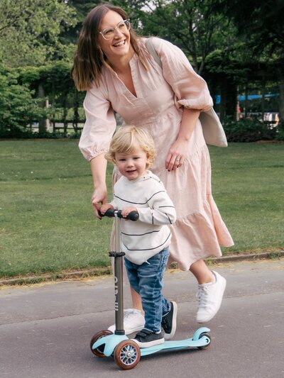 fun candid family picture showing a mom helping her toddler play on a scooter. they both smile. taken in Christchurch Botanic Gardens in New Zealand.