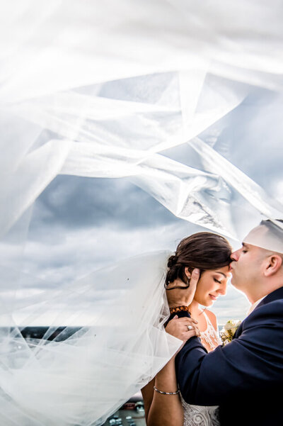 groom kissing bride on forehead while veil blows overhead