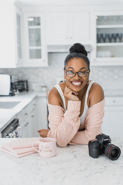 Houston Wedding Photographer, Joy Henderson. Standing in a kitchen. Leaning on the counter surrounded by a pink coffee mug, pink notebooks, and her Canon DSLR Camera. She is wearing glasses, a white tank top, and a pink cardigan.