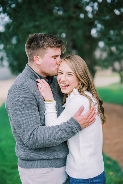 Man kissing woman on the head while she holds up her engagement ring on her hand