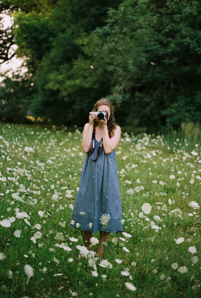 Heather, of Among the Oaks, stands amidst wild flowers and takes a photo, representing her film wedding photography offerings. 