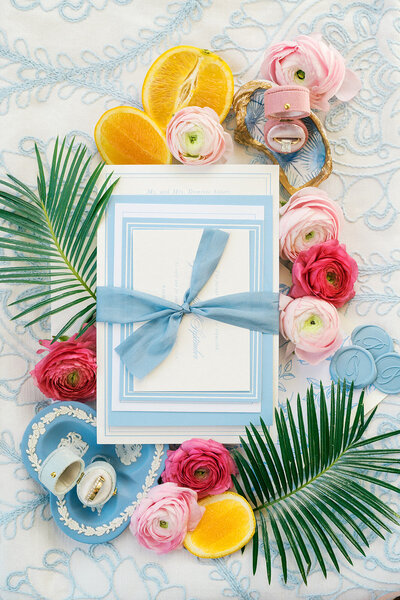 Blue and white wedding invitation suite is surrounded by pink flowers and palm leaves for coastal wedding