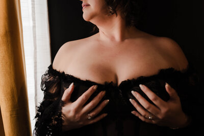 A woman in black lingerie holds her breasts while looking out a window