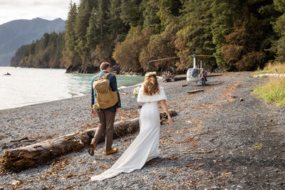 A bride and groom walk along a remote Alaskan beach toward a waiting helicopter, surrounded by rugged coastline and dense evergreen forests. The groom carries a backpack, and the bride’s wedding dress flows behind her, capturing the essence of an adventurous elopement in the Alaskan wilderness.