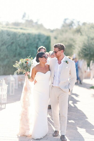 Newlywed couple walk arm in arm through a sunlit path surrounded by olive trees at a private villa estate in Lefkada