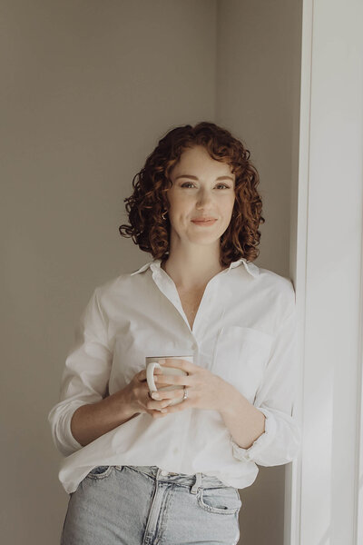 Woman leaning against the wall, holding a coffee cup with both hands and looking out the window