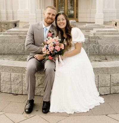 bride and groom sitting out side of LDS temple holding light pink orange and blue bouquet