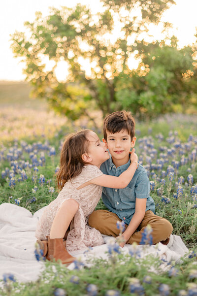 A girl hugs her brother and kisses his cheek in the glowing light of golden hour. They sit on a blanket in a field of San Antonio bluebonnets.