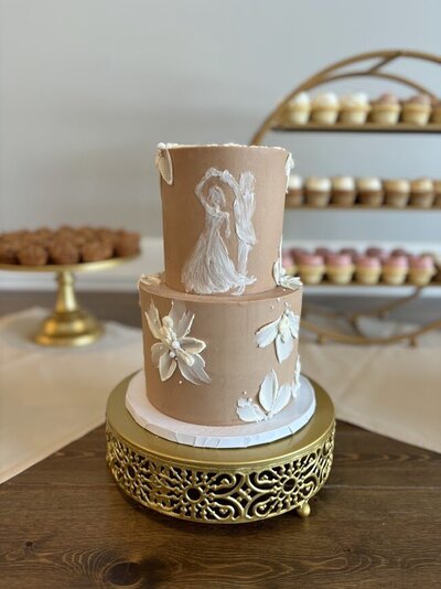 A two tier cake sits on a gold cake stand, in front of other gold cake stands with mini cupcakes and tarts (background). The cake has brown chocolate frosting with a white silhouette of a couple dancing together and white buttercream flowers. 