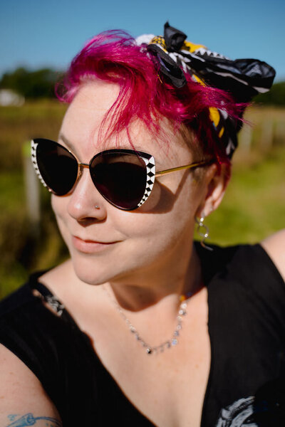 A person with pink hair and large sunglasses slightly smiling while standing outside.