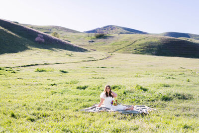 Girl sitting next to bushes and flowers touching her hair and looking off into the distance.