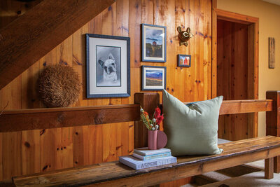 A cozy hallway in a wood cabin with wood-paneled walls decorated with framed artwork, including a portrait of a dog, and a rustic wooden animal head. A wooden bench sits along the wall with a light green pillow, a stack of books (one titled "Aspen Style"), and a pink vase with dried flowers. A textured brown decorative object rests on a wooden shelf above the bench. A doorway leading to another room is visible in the background.