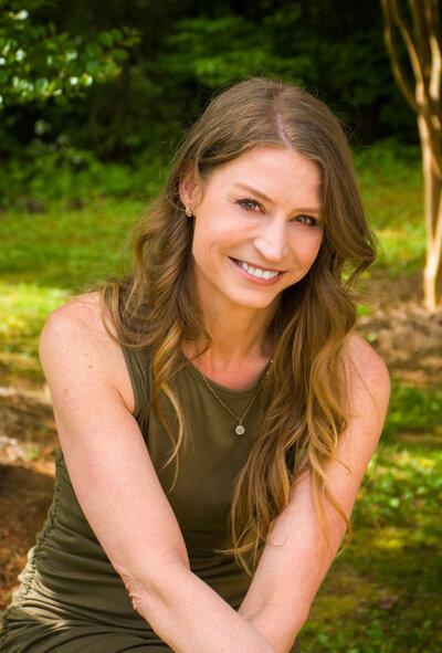 A woman with long brown hair smiles while sitting outdoors. She is wearing a sleeveless olive green top and a necklace. The background is lush with greenery, including trees and grass.
