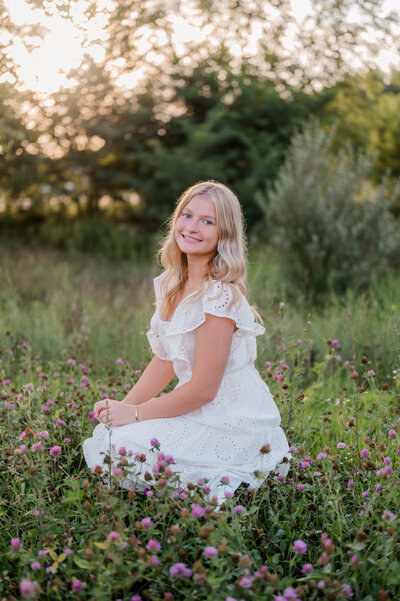 Girl in white dress kneels in field with purple flowers.