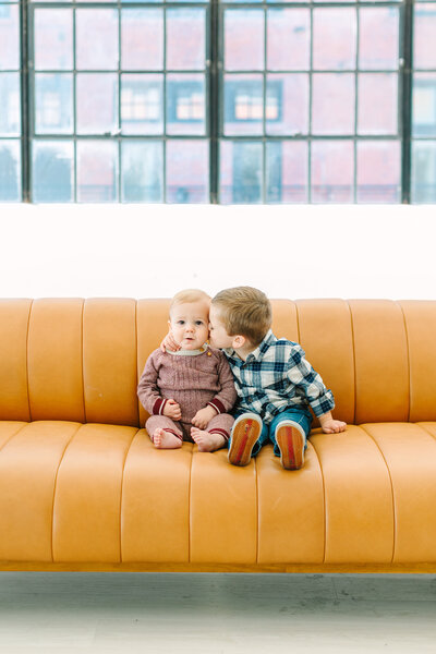 Little boy kissing baby on the cheek while sitting on a yellow couch