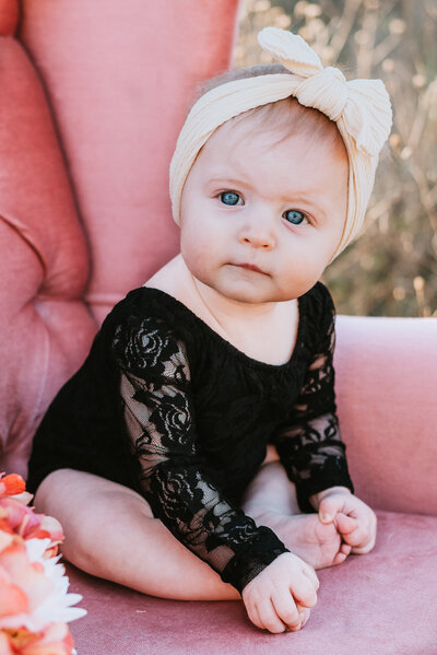 Baby girl in black onsie sitting in pink chair for six month photos..