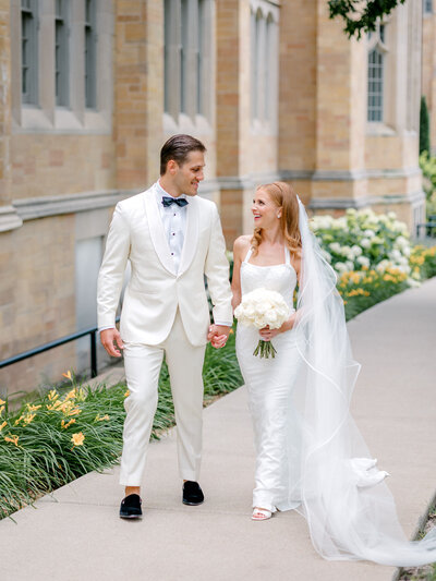 Bride with a cathedral veil holds hands with groom in a white tux walking along a sidewalk with church facade behind them
