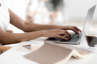 Women's hands working on SEO strategy on a laptop sitting at a table with a journal and cup of coffee.