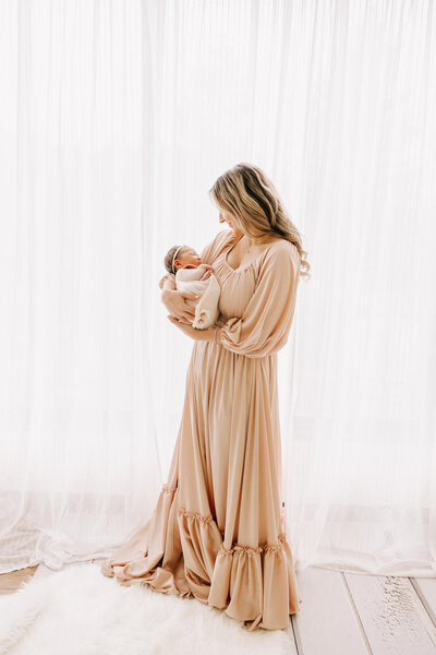 stunning portrait of a mom standing in front of a white set of curtains over the window. She is wearing a long blush dress and holding her baby girl