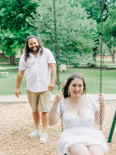 A man pushes a smiling woman on a swing at a park. Both are dressed in light-colored summer clothes, with trees in the background. Captured by a San Antonio Wedding Photographer, they appear happy and relaxed.