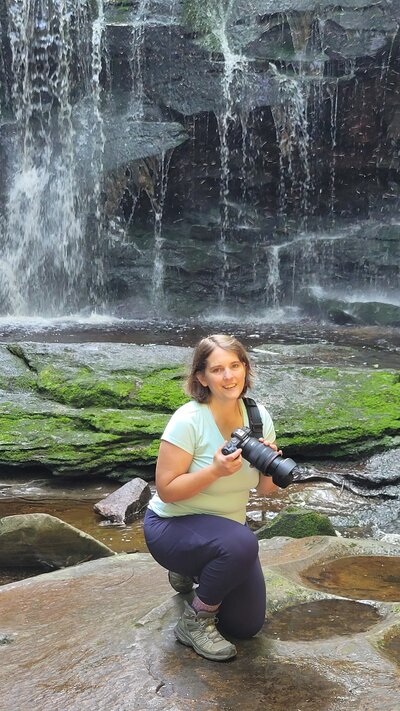 Joy Granados holding her camera while sitting on rocks near a waterfall, captured behind the scenes as Northern Virginia family photographer of Capture Joy Photography.