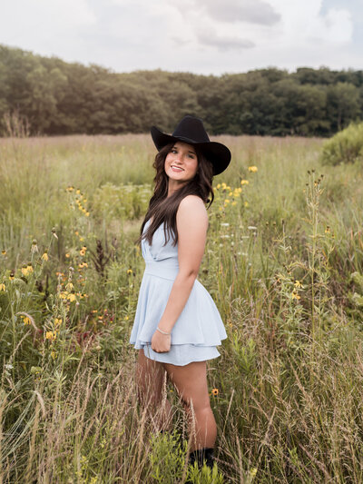 high school senior girl in blue dress and black cowboy hat posing in field for senior portraits