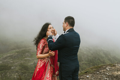 groom brushing tears off his brides face during their alaska elopement in whittier, with green rolling mountains, she is wearing a red sari.