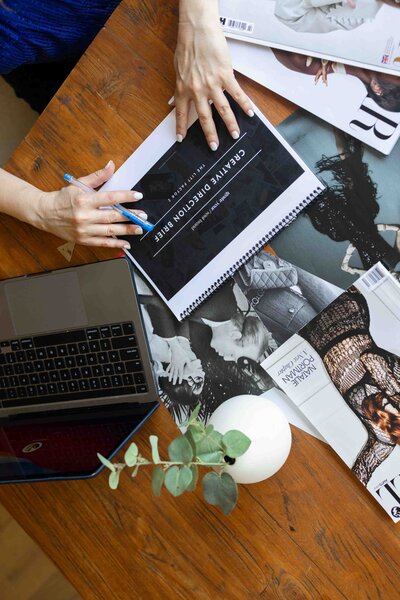 Creative direction brief and laptop on wooden desk with editorial magazine spreads and eucalyptus styling during brand strategy session