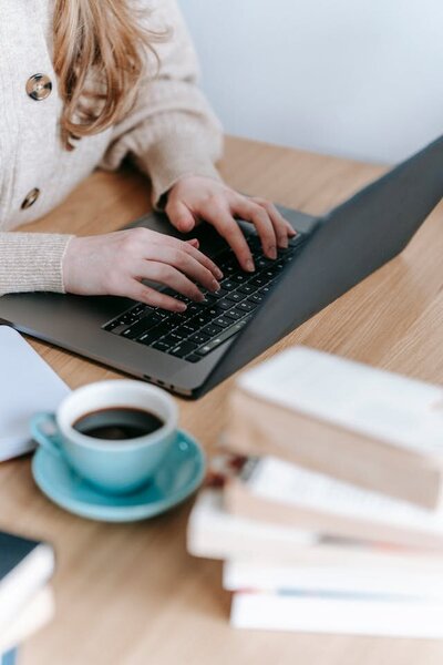 Gros plan sur une jeune femme qui tape à l'ordinateur sur son bureau, avec un café et des livres à côté d'elle