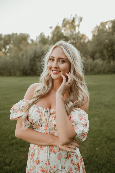 Senior portrait session by Bailee Freeman Photography for a blonde model with long hair. The model is wearing a pink and cream floral dress against a field of green grass and green trees in the distance. 