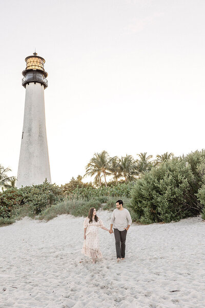Parents to be walking on the beach with a lighthouse during their maternity session